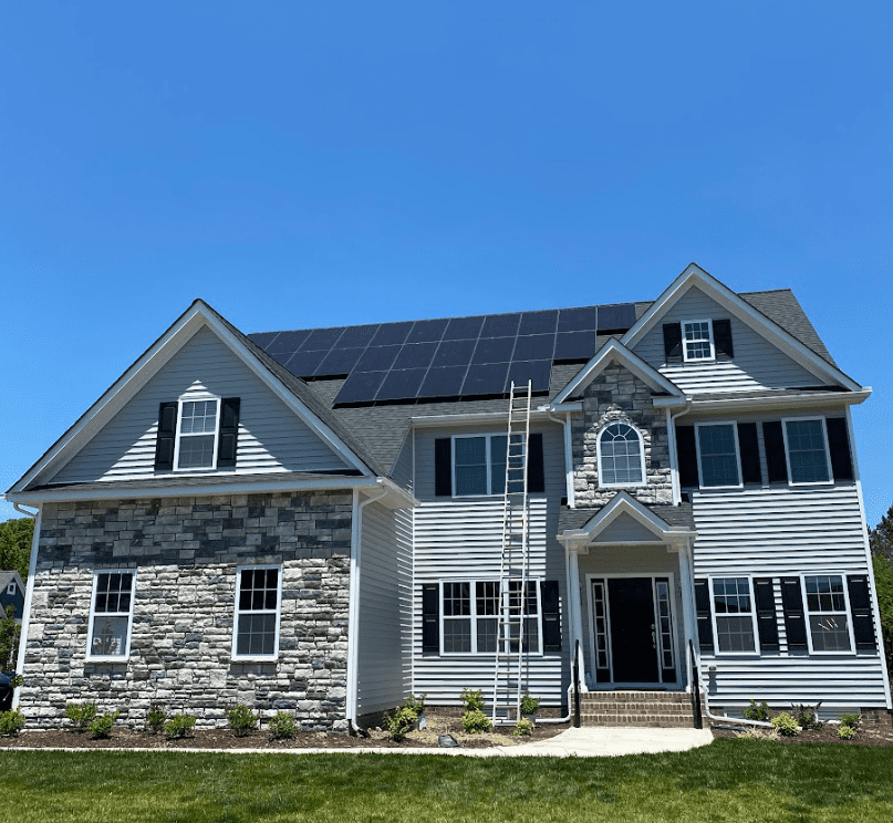 A two-story house with gray siding and stone accents, featuring solar panels on the roof. A ladder leans against the entrance under a clear blue sky.